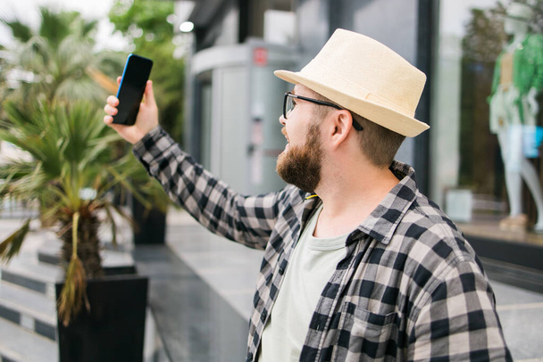 Brodaty człowiek robi selfie poprzez spacery smartfona w centrum miasta stanowi przed nowoczesnym budynku miasta cieszy się czas wolny. Miejski styl życia i technologia - Zdjęcie, obraz