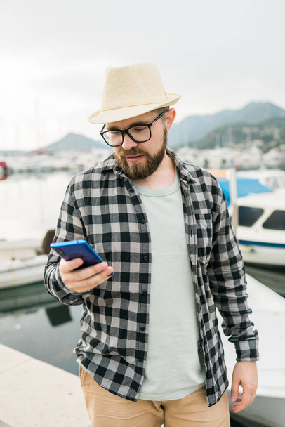 Handsome man tourist check in social networks during summer walk among sea marina with boats and yacht background - traveller and social media app - Zdjęcie, obraz