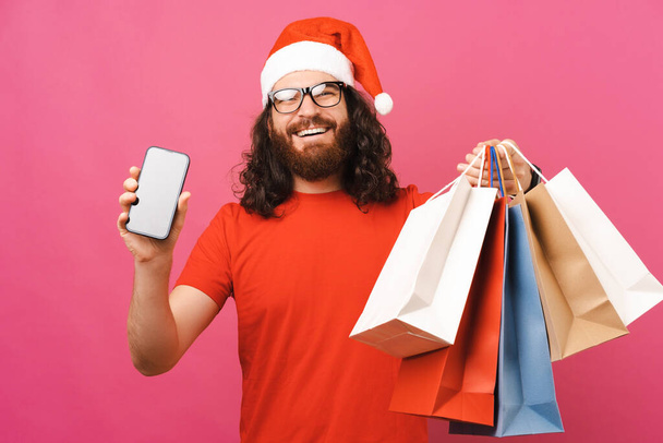 Hombre extático en sombrero de Navidad muestra una pantalla del teléfono mientras sostiene bolsas de compras. Estudio sobre fondo rosa. - Foto, imagen