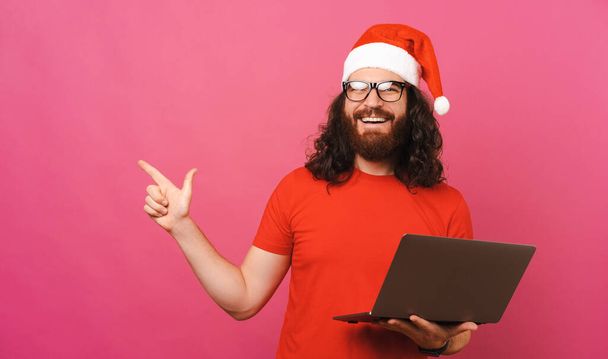 Happy man wearing Christmas hat is pointing aside and holding a laptop. Studio shot over pink background. - Photo, Image