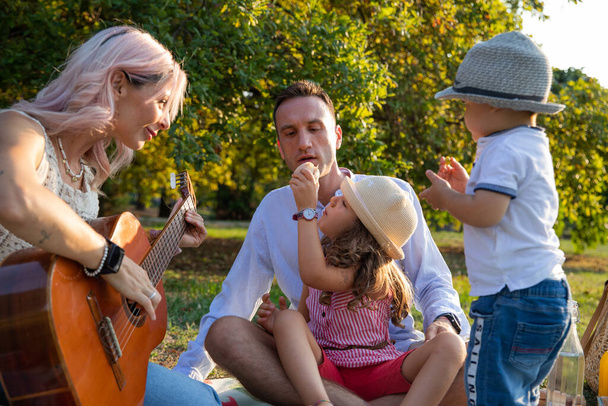 Família de quatro com crianças pequenas no parque piquenique enquanto a mãe toca guitarra. - Foto, Imagem