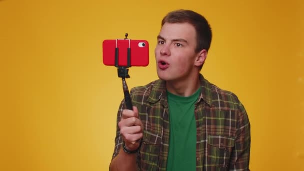 Vay canına. Etkileyici blog yazarı cep telefonu özçekim çubuğuyla selfie çekiyor abonelerle online video görüşmesi yapıyor. 20 'li yaşlarda genç bir çocuk sarı stüdyo duvarında tek başına. - Video, Çekim