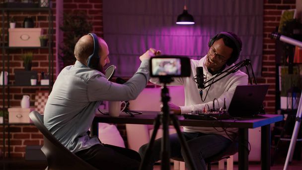 Static tripod shot of internet radio host recording podcast using digital video camera greeting guest in home studio. African american podcaster doing fist bump in interview with famous influencer. - Foto, imagen