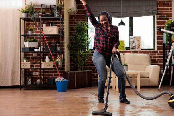 Authentic woman using modern vacuum cleaner listening to favorite music with wireless headphones. Housewife having fun while finishing spring cleaning at home, good vibes and positive energy. - Фото, зображення