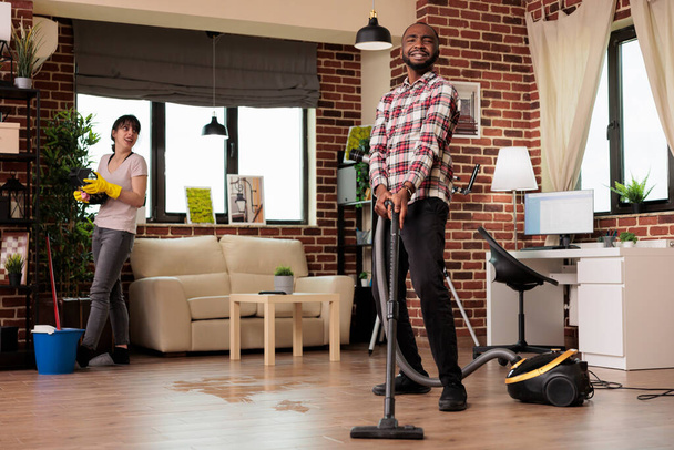 Multiracial couple happily cleaning at home, african american man vacuuming the whole place while woman dusts the shelves. Working as a team to keep house clean and tidy. - Photo, Image