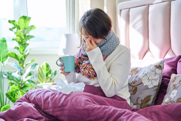 Sick female at home in bed under blanket with handkerchiefs. Middle-aged woman with scarf, mug of hot drink, with symptoms of cold. Flu season, autumn winter respiratory diseases - Photo, Image