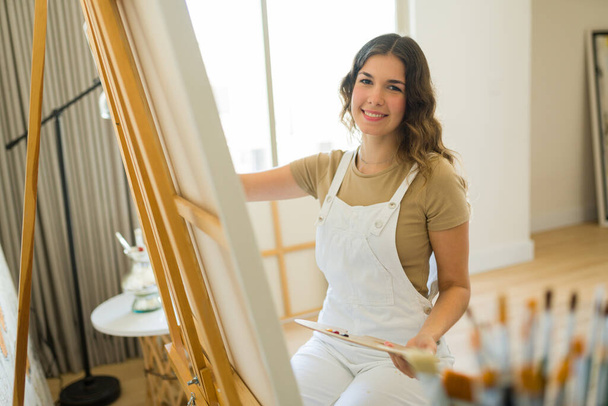 Portrait of a happy young woman artist smiling using a paintbrush and working on a new painting  - Photo, Image