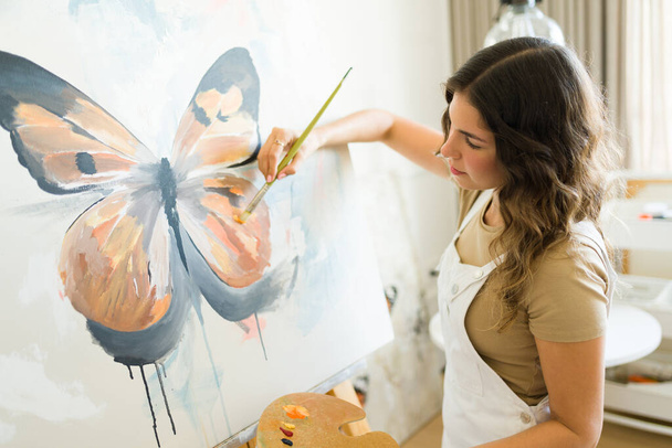Γυναίκα εμπνευσμένη καλλιτέχνης γυναίκα χρησιμοποιώντας ένα πινέλο και ζωγραφική μια πεταλούδα σε ένα καμβά, ενώ δημιουργώντας τέχνη - Φωτογραφία, εικόνα