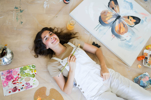 Vista dall'alto di un'attraente pittrice creativa che si rilassa sul pavimento e tiene un pennello sorridendo accanto al suo dipinto - Foto, immagini