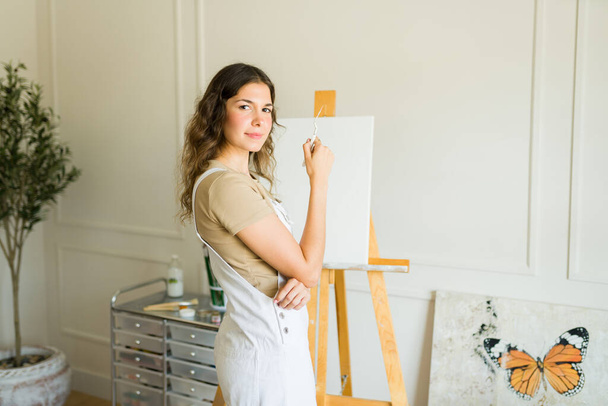 Jovem artista feminina bonita sentindo criativo segurando um pincel de pé na frente de uma tela em branco no cavalete - Foto, Imagem
