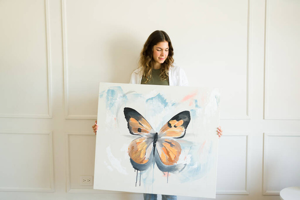 Gyönyörű kaukázusi fiatal nő művész mutatja neki kész kreatív festmény előtt egy fehér fal - Fotó, kép