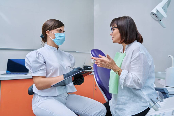 Dentist and woman patient in dental clinic, doctor nurse writing on clipboard consulting, middle aged female talking about disturbing dental problem. Dentistry, treatment, dental health care concept - Fotoğraf, Görsel