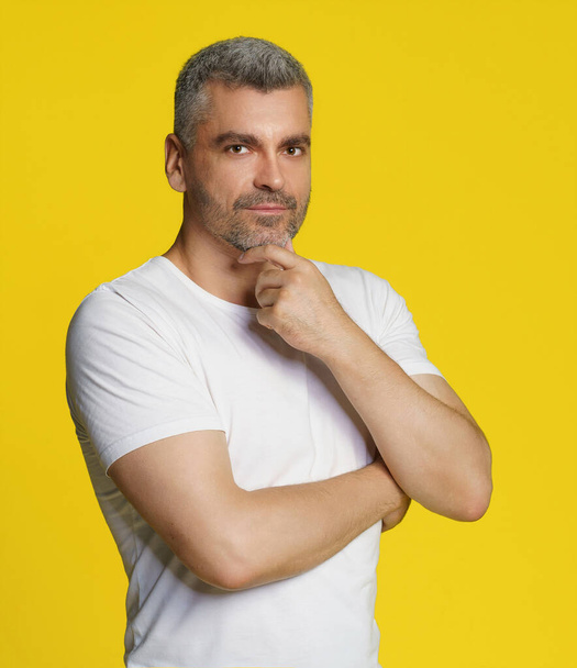 Middle aged grey hair athletic man posing with hands crossed touching chin wearing white t-shirt isolated on yellow background. Thoughtful fit, muscular middle aged man. - Foto, afbeelding