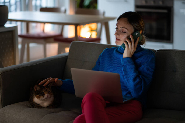Kobieta freelancer rozmawia na smartfonie z kolegą o niezbędnych szczegółach w projekcie i głaszcze kota domowego leżącego na wygodnej sofie. Młoda businesswoman pracuje w pół ciemnym mieszkaniu - Zdjęcie, obraz