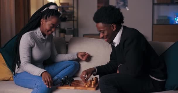 Вечером после занятий двое студентов африканского происхождения сидят на диване в гостиной и играют в шахматы. Девочка и мальчик делают свои движения, болтают, улыбаются и веселятся. - Кадры, видео