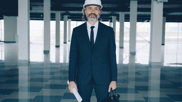 Portrait of experienced chief engineer holding blueprint and wearing helmet standing in modern plant building alone ready for real estate deal - Fotoğraf, Görsel