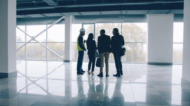 Ομάδα μηχανικών άνδρες και γυναίκες συζητούν νέο κτίριο εσωτερικό έλεγχο σχέδιο και μιλώντας στέκεται πάνω από πανοραμική φόντο παράθυρο - Φωτογραφία, εικόνα