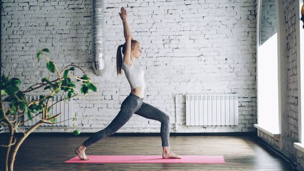 Young slim blond girl is doing yoga complex in nice studio with white walls. She is starting with warrior poses, then bending forward and backward and finishing with namaste. - Photo, image