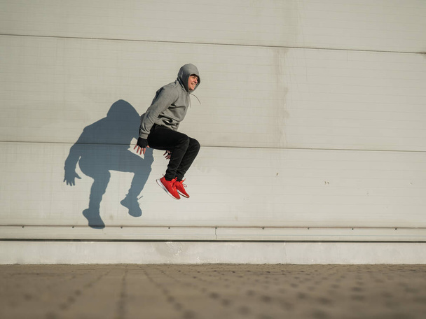 Caucasian man in hoodie jumping high with hands on knees outdoors - Zdjęcie, obraz