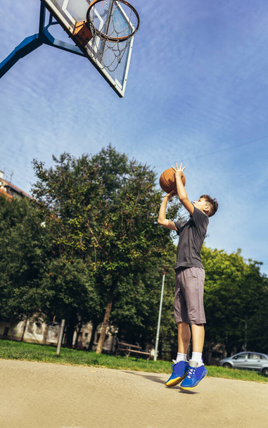 Χαριτωμένο αγόρι με γκρι μπλουζάκι παίζει μπάσκετ στην παιδική χαρά της πόλης. - Φωτογραφία, εικόνα