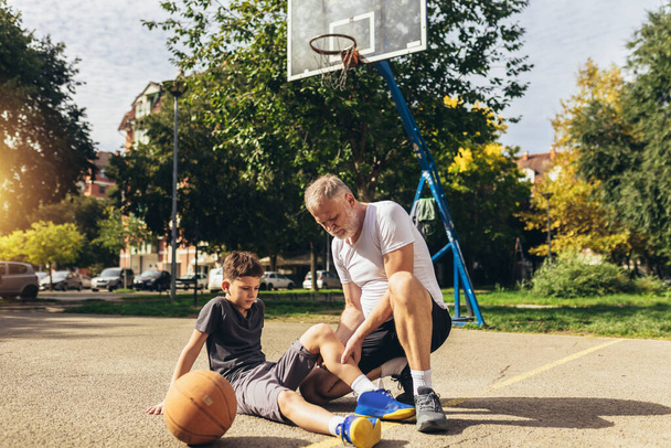 Padre ayudando a niño con trauma en las piernas después de jugar baloncesto. Lesiones deportivas.  - Foto, Imagen