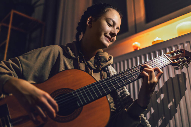 Дівчина-підліток сидить під ковдрою біля опалювального радіатора зі свічками та грає на гітарі.
 - Фото, зображення