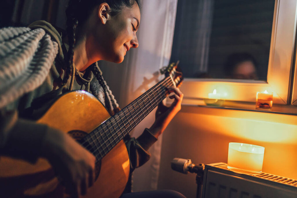 Nastolatka siedzi pod kocem w pobliżu grzejnika ze świecami i grać na gitarze. Rosnące koszty w prywatnych gospodarstwach domowych za rachunek za gaz z powodu inflacji i wojny, kryzys energetyczny - Zdjęcie, obraz