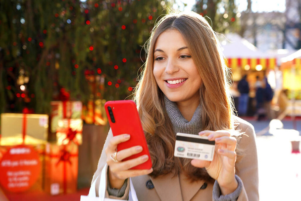 Приваблива молода жінка платить свої подарунки онлайн за допомогою смартфона і кредитної картки з різдвяними ринками і деревом з коробками на задньому плані. - Фото, зображення