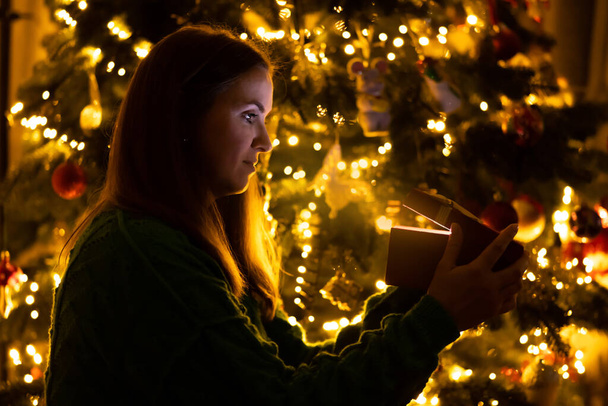 Happy beautiful woman in front of Christmas tree opening gift box and looking at the magic light inside the box. Marry Christmas and Happy New Year concept - Foto, Imagen