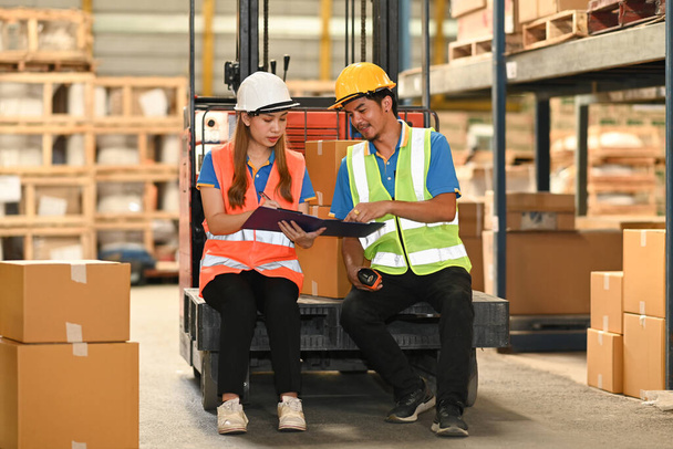 Two warehouse workers wearing safety hard hat and vest sitting on forklift truck in retail warehouse checking newly arrived goods. - Φωτογραφία, εικόνα