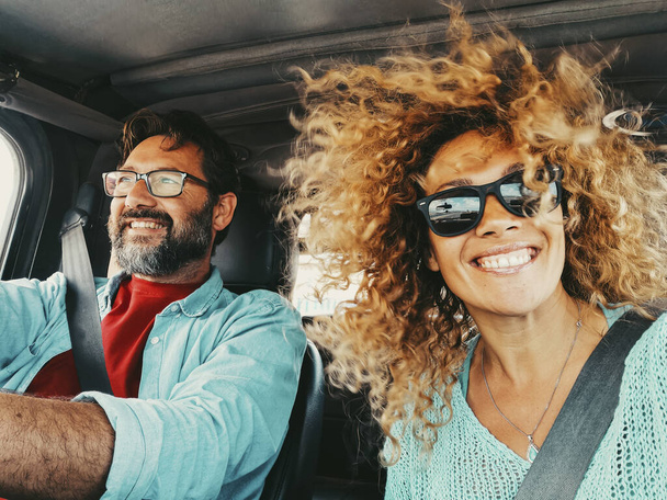 Happy young couple traveling by car together having fun. Woman taking selfie picture in road trip with her husband driving the vehicle. Transportation. People with safety belt inside car enjoying - Photo, Image