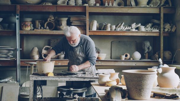 Hardworking silver-haired man is working with clay on potters wheel in workplace, shaping piece of loam. Beautiful ceramic utensils, handmade pots and vases on shelves are visible. - Foto, Imagem