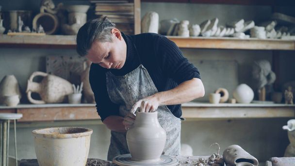 Experienced male ceramist is forming ceramic vase from clay on spinning throwing wheel while working in cozy potters workshop. Professional is focused on work. - Zdjęcie, obraz