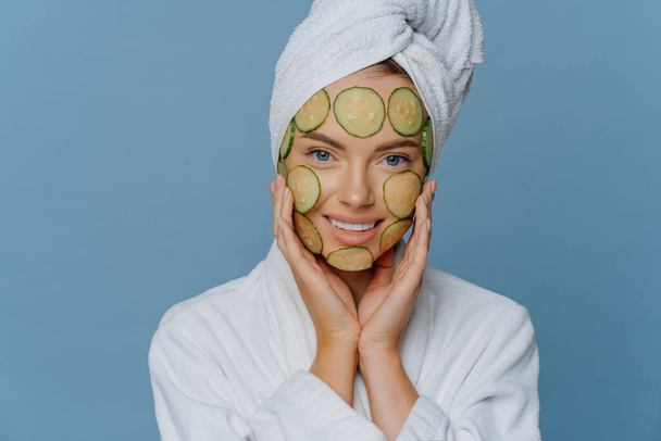 Young beautiful woman applies cucumber slices on face has perfect clean skin touches cheeks gently dressed in comfortable dressing gown has towel wrapped on head. Cosmetology and spa concept - Photo, image