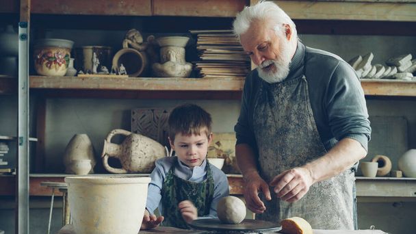 Заботливый седовласый дедушка учит юного симпатичного внука работать с глиной на метательном колесе в маленькой уютной мастерской. Концепция керамики, семейного хобби и ремесла
. - Фото, изображение