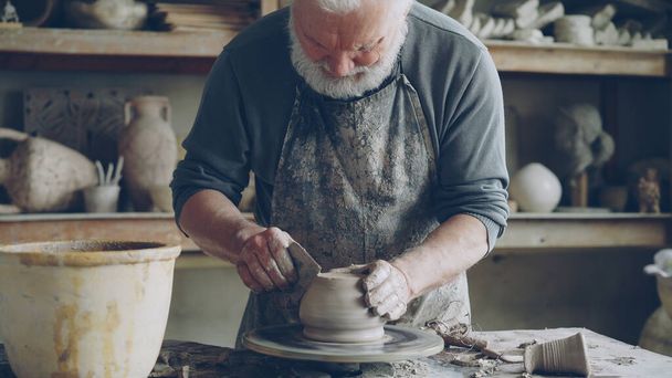 Senior male potter is working at bottom part of ceramic pot on spinning potters wheel in workplace. Creating traditional eathenware and conventional pottery concept. - Zdjęcie, obraz