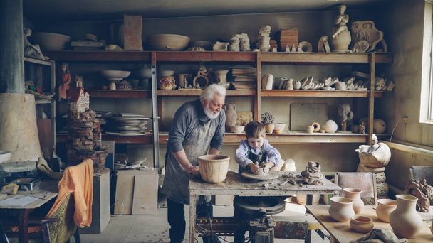 Experienced potter loving prandparent is teaching his young grandchild pottery in small home workshop. Boy is working with throwing wheel, grandpa is talking to him. - Fotoğraf, Görsel
