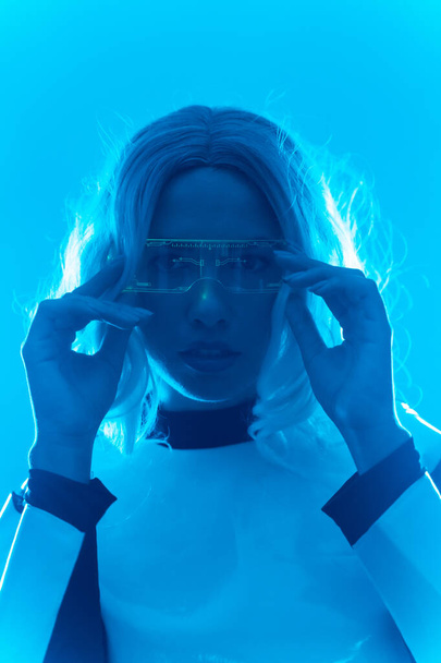 Young woman with futuristic suit and glasses with blue led lights, virtual or metaverse concept, looking at camera - 写真・画像