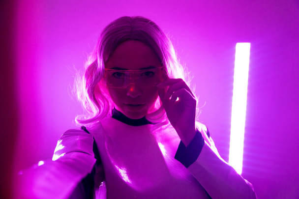 Woman in futuristic suit and glasses with pink led lights, virtual concept, metaverse, selfie with camera - Photo, Image