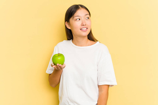Junge Asiatin isst einen Apfel isoliert auf gelbem Hintergrund und blickt lächelnd, fröhlich und angenehm zur Seite. - Foto, Bild