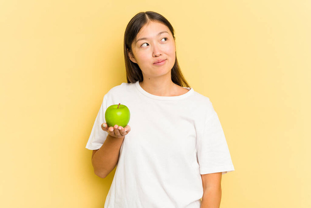 若いアジアの女性は、目標と目的を達成することを夢見て黄色の背景に隔離されたリンゴを食べる - 写真・画像