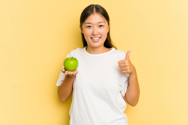 Junge Asiatin isst einen Apfel isoliert auf gelbem Hintergrund, lächelt und hebt den Daumen - Foto, Bild