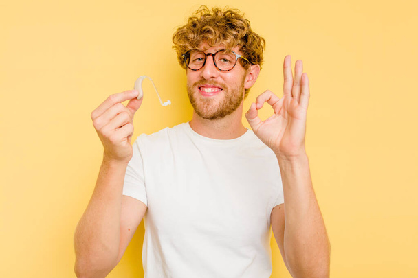 Young caucasian man wearing hearing aid isolated on yellow background cheerful and confident showing ok gesture. - Fotografie, Obrázek