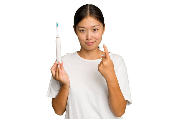 Young Asian woman holding an electric toothbrush isolated on green chroma background pointing with finger at you as if inviting come closer. - Photo, Image