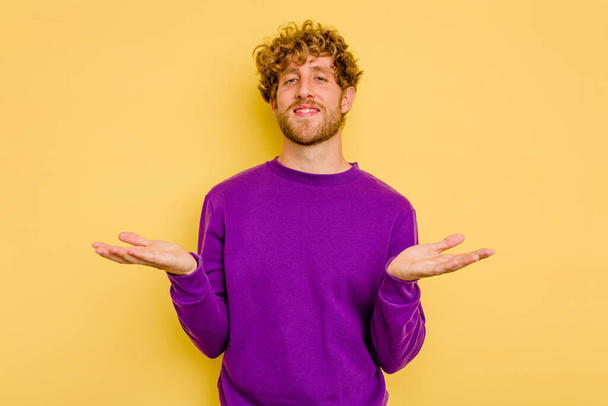 Young caucasian man isolated on yellow background makes scale with arms, feels happy and confident. - Photo, Image