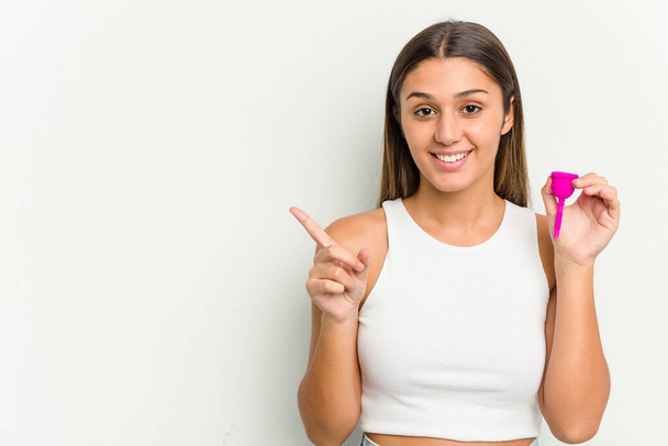Jonge vrouw met een menstruatiecup geïsoleerd op witte achtergrond glimlachend en wijzend opzij, toont iets op lege ruimte. - Foto, afbeelding