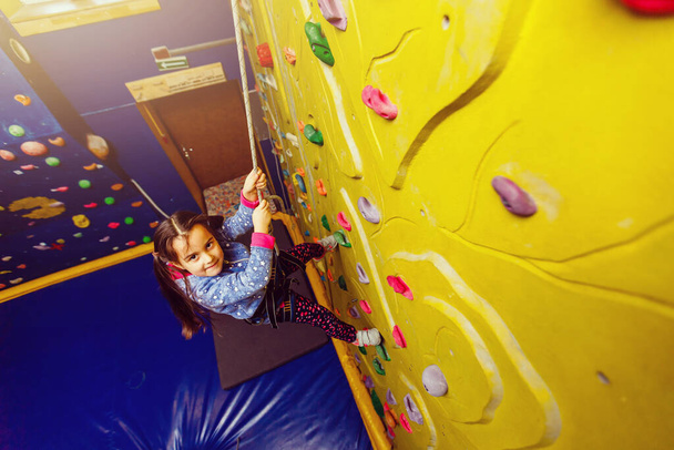 Little baby girl with funny hear style climbing vertical wall and man belaying her from below - Photo, Image