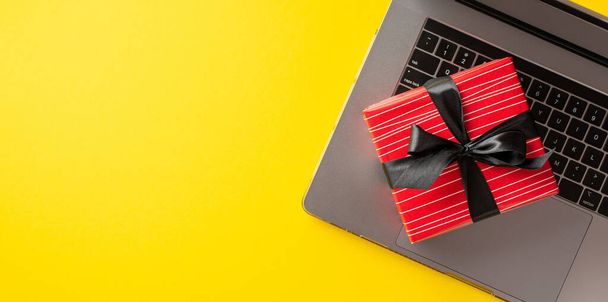 Διαδικτυακή ιδέα Δευτέρας. Top view φωτογραφία του trendy κόκκινο giftbox με κορδέλα τόξο στο φορητό υπολογιστή σε απομονωμένο κίτρινο φόντο με copyspace - Φωτογραφία, εικόνα
