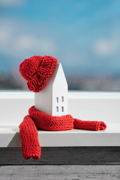 Miniaturowy dom w czerwonym szaliku i kapeluszu na parapecie. Koncepcja pasywnego ogrzewania domu. Izolacja cieplna budynku lub mieszkania. Kryzys. - Zdjęcie, obraz