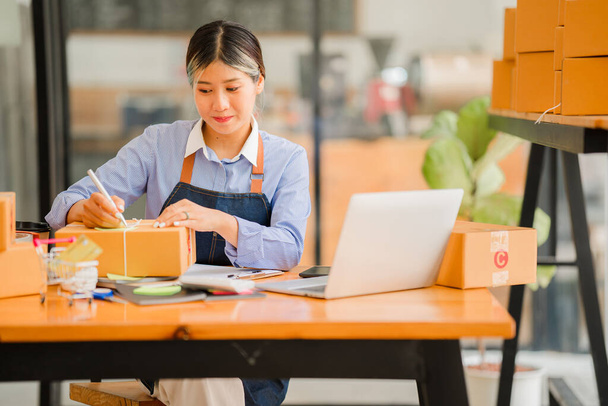 Small business start-up, SME, owner of successful young Asian woman entrepreneurs smiling. use a smartphone or laptop Receive and check online orders to prepare packing boxes. selling ideas online - Photo, Image